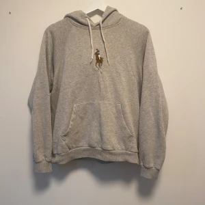 Grå hoodie från Polo Ralph Lauren - Snygg grå hoodie från Polo Ralph Lauren med klassisk broderad polospelare på bröstet. Tröjan har huva med snörning och en stor magficka. Perfekt för dig som gillar en clean och sportig stil.