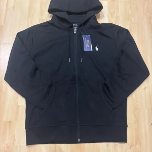 Svart hoodie från Polo Ralph Lauren - Svart hoodie från Polo Ralph Lauren med dragkedja framtill och klassisk huva. Tröjan har två fickor, snörning vid huvan och den ikoniska vita loggan broderad på bröstet. Perfekt för en chill och stilren look.