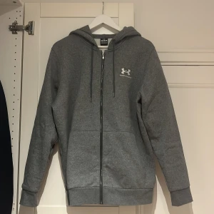 Grå hoodie från Under Armour  - Säljer en grå hoodie från Under Armour i storlek M. Tröjan har dragkedja, huva med snörning och två fickor framtill. Perfekt för chill dagar eller träning. Under Armour-logga broderad på bröstet. Mjuk insida och klassisk passform.