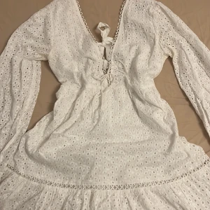 Vit broderad kortklänning med knyt - Superfin vit kortklänning i bomull med broderade detaljer och volangkant. Klänningen har långärmade puffärmar, v-ringning med snörning framtill och knyt i ryggen. Perfekt för skolavslutning eller student 