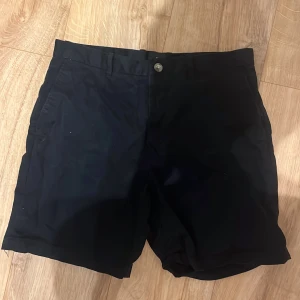 Svarta shorts från H&M Regular Fit - Klassiska svarta shorts från H&M Regular Fit med normal passform. De har bälteshällor, knäppning fram och två bakfickor med knapp. Perfekta för sommaren och enkla att matcha med allt. Materialet är bomull och polyester för en skön känsla.