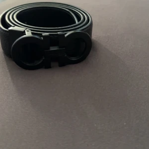 Svart Ferragamo skärp i skinn - Snyggt svart skärp från Ferragamo med ikoniskt dubbel-Gancini-spänne i matt svart metall. Bältet är gjort i äkta skinn med en stilren och klassisk design. Perfekt accessoar för att lyfta din outfit med en lyxig touch. 120 cm