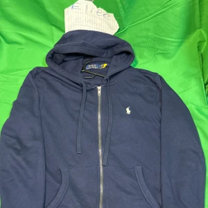 Marinblå hoodie från Polo Ralph Lauren - Snygg marinblå hoodie från Polo Ralph Lauren med dragkedja framtill, huva med snörning och klassisk vit logga på bröstet. Mjuk bomullskvalitet och två fickor framtill. Perfekt för chill dagar eller när du vill ha en clean streetstyle.Liknande modell som originalet!