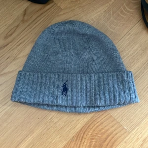 Grå stickad mössa från Polo Ralph Lauren - Snygg grå stickad mössa från Polo Ralph Lauren med klassisk blå broderad logga framtill. Mössan har bred ribbad uppvikning och är perfekt för kyliga dagar. Enkel och stilren design som passar till det mesta. PRIS KAN DISKUTERAS 🙂