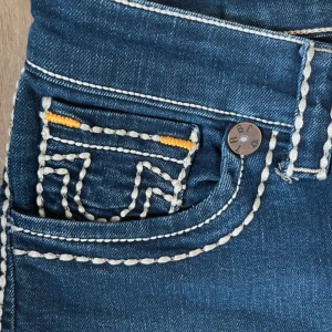 True Religion Joey jeans med kontrastsömmar - Säljer ett par blå True Religion Joey jeans med ikoniska vita kontrastsömmar och detaljer i gult. Klassisk femficksmodell med tydlig logga bak och markerade bakfickor. Jeansen har lite utsvängda och är tillverkade i kraftigt denimtyg.en ersättnings knapp där framme
