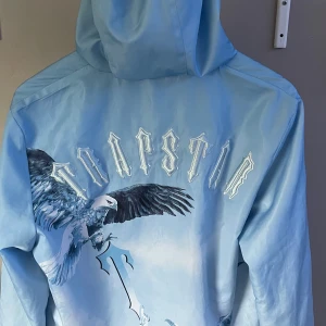 Trapstar vindjacka  - Säljer en ljusblå vindjacka från Trapstar med huva och dragkedja. Jackan har ett stort tryck av en örn på både fram- och baksidan samt Trapstar-logga broderad på ryggen. Elastiska muddar vid ärmslut och två fickor framtill. Perfekt för dig som vill sticka ut. Säljer pga att den inte passar längre. Storlek S men passar även xs , ändå dåliga är att ärmen är invikt men det går säkert att fixa