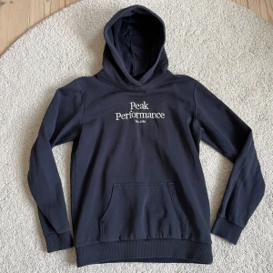 Peak Performance hoddie - Snygg mörk blå hoodie från Peak Performance med vit broderad logga på bröstet. Klassisk känguruficka framtill och huva. Storlek 170/S