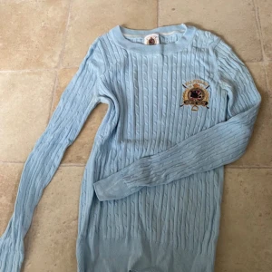 Ljusblå kabelstickad tröja falsterbo hs  - Kabelstickad ljusblå tröja från Gina Tricot med broderad Falsterbo Horse Show-logga på bröstet. Tröjan har rund halsringning och långa ärmar, ridinspirerade detaljer. endast en liten defekt, se bild 