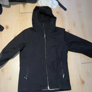 Svart vindjacka Peak Performance M - Svart vindjacka från Peak Performance med huva och hel dragkedja framtill. Jackan har två snedställda fickor med dragkedja och en stilren, enkel design. Perfekt för blåsiga dagar och passar till många olika outfits.