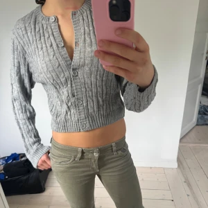 Grå croppad kofta med knappar - Säljer en grå croppad kofta med kabelstickat mönster och rund hals. Koftan har långa ärmar och knäppning framtill med flera knappar.