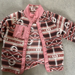 Mönstrad teddyjacka i rosa och brunt - Säljer en riktigt mysig teddyjacka med unikt mönster i rosa, brunt och vitt. Jackan har hög krage, tryckknappar och en praktisk bröstficka med dragkedja. Perfekt för dig som vill sticka ut med en cool och färgglad look.