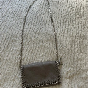 Grå  axelväska från Tiamo med kedja - Snygg grå axelväska från Tiamo med grov kedja som axelrem och flätad kant i metall. Väskan är i skinnimitation med struktur och har ett fack med dragkedja inuti. Perfekt storlek för det viktigaste och ger en cool vibe till din outfit.