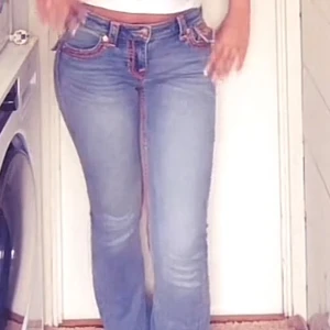 True Religion jeans med rosa sömmar - Säljer ett par blå True Religion jeans med ikoniska rosa kontrastsömmar. Jeansen har klassiska fickor med lock och knapp, samt tydlig logopatch bak. Modellen är rak med normal passform och tillverkad i jeansmaterial.