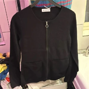 Svart kofta med dragkedja från Wera - Svart kofta från Wera med rund halsringning, hel dragkedja framtill och två stora fickor. Långärmad och i mjukt, stickat material. Perfekt för lager-på-lager och enkel att matcha med allt. Helt ny med lappar