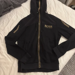 Svart hoodie från Hugo Boss med guld - Svart hoodie från Hugo Boss med gulddetaljer och dragkedja framtill. Broderad BOSS-logga på bröstet, huva med snörning och coola guldstreck på ärmarna. Mjuk bomullsmix och ribbade muddar. Perfekt för en stilren och sportig look.