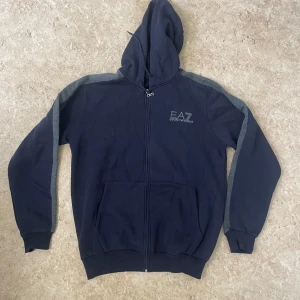 Marinblå EA7 hoodie med grå detaljer - Snygg marinblå hoodie från EA7 Emporio Armani med grå ränder längs ärmarna. Tröjan har huva, dragkedja framtill och två stora fickor. Perfekt för en chill och sportig look.