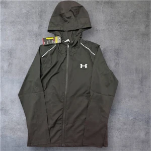 Svart zip från Under Armour - Svart vindjacka från Under Armour med huva och hel dragkedja framtill. Jackan har vita detaljer vid axlarna och logga på bröstet samt stor Under Armour-text på ryggen. Tillverkad i lätt polyester, perfekt för aktiva dagar utomhus.