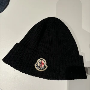 Svart ribbad Moncler mössa - Svart ribbstickad mössa från Moncler med klassisk uppvikt kant och broderad logotyp framtill. Snygg och enkel design som ger en clean look, perfekt för dig som gillar exklusiva märken. Materialet är mjuk ull som håller dig varm under kalla dagar.