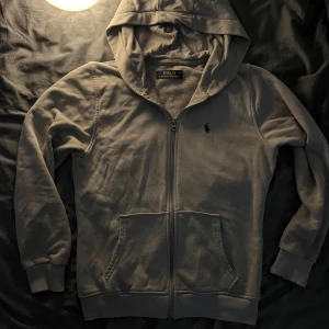 Grå hoodie från Polo Ralph Lauren - Säljer en stilren grå hoodie från Polo Ralph Lauren med dragkedja, huva och två fickor framtill. Klassisk broderad logga på bröstet och mjukt innerfoder. Perfekt för dig som gillar enkel och clean streetstyle.