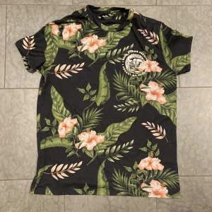 Svart blommig t-shirt från Bershka XS - Svart t-shirt från Bershka i storlek XS med tropiskt blommönster i grönt och rosa. Tryck med texten 'Lost Paradise' och illustration av strandhus på ryggen. Skön passform och perfekt för sommarvibbar. Materialet är mjukt och känns som bomull.