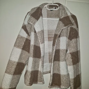Rutig teddyjacka i beige och vitt - Mysig tunnare teddyjacka med stora rutor i beige och vitt. Jackan har en klassisk krage och är tillverkad i teddy-material som ger en cool och avslappnad vibe. Perfekt för dig som gillar trendiga och bekväma plagg med lite extra attityd. Passar en M oxå då den är lite oversized! Går inte att stänga.