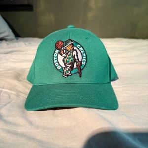 Grön Boston Celtics keps Mitchell & Ness - Säljer en grön snapback-keps från Mitchell & Ness med Boston Celtics broderad logga framtill. Kepsen har justerbar plastrem bak och vit broderad text. Materialet är bomull och designen är klassisk med böjd skärm. Perfekt för dig som gillar basket och streetwear.