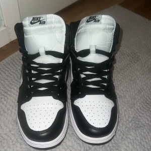 Nike Air Jordan 1 svartvita sneakers - Säljer ett par Nike Air Jordan 1 i svart och vitt. Klassisk high-top modell med svart swoosh, vita paneler och svarta detaljer. Skorna har snörning, rund tå och är tillverkade i skinn. Perfekta för dig som vill ha en ikonisk och stilren sneaker.