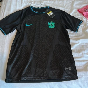 Svart Brasilien Nike fotbollströja S - Säljer en svart Brasilien fotbollströja från Nike med Dri-Fit material. Tröjan har korta ärmar, turkosa detaljer vid krage och ärmslut samt ett mönstrat parti framtill. Brasilien-emblemet i neonfärg och Nike-logga på bröstet. Perfekt för dig som älskar fotboll och snygg design.