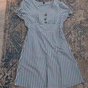 Blåvit randig kortklänning med knappar - Säljer en söt blåvit randig kortklänning med v-ringning och tre dekorativa knappar framtill. Klänningen har puffiga korta ärmar och ett markerat midjeparti som ger en fin siluett. Perfekt för dig som gillar fräscha och somriga vibes!