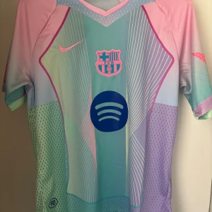 Barca rosa och blå tröja - Unik FC Barcelona fotbollströja från Nike i pastellfärger med klubbmärke och Spotify-logga på bröstet. Tröjan har korta ärmar, grafiskt mönster i rosa, blått, lila och mintgrönt samt diskreta ränder och prickar. Tillverkad i lätt och ventilerande funktionsmaterial.