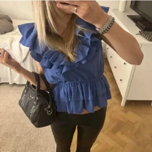 volangblus - Supersöt blå blus med volanger och v-ringning. Ärmlös modell med resår i midjan som ger en snygg passform. Luftigt tyg, perfekt för varmare dagar. Volangerna ger en trendig och lekfull look. Priset kan diskuteras