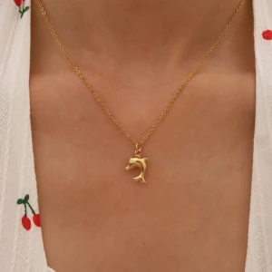 Guldigt halsband med delfinberlock - Supersött halsband i guldton med en detaljrik delfinberlock. Kedjan är tunn och elegant, perfekt för att ge en lekfull touch till din stil. Delfinen har en mjukt böjd form och glänsande finish som fångar ljuset snyggt. Rostfritt 18k guldpläterad 