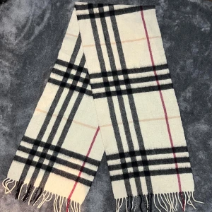 Rutig halsduk från Burberry i ull - Klassisk halsduk från Burberry i mjuk ull med ikoniskt rutigt mönster i vitt, svart, beige och röda detaljer. Långa fransar i ändarna och tidlös design som lyfter vilken outfit som helst.