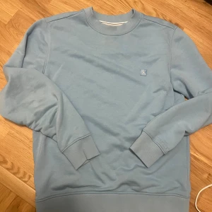 Ljusblå sweatshirt från Calvin Klein - Säljer en stilren ljusblå sweatshirt från Calvin Klein Jeans med broderad CK-logga på bröstet. Tröjan har rund hals, ribbade muddar och är tillverkad i mjuk bomull. Perfekt för en chill och snygg look.