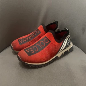 Röda Dolce & Gabbana sneakers - Säljer ett par röda Dolce & Gabbana sneakers med svart och vit logga på ovansidan och hälen. Skorna har en stickad ovandel i syntetmaterial, elastisk passform och grov sula för extra attityd. Perfekta för dig som vill sticka ut med en sportig och trendig look.