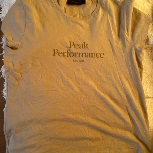 Beige Peak Performance t-shirt - Snygg beige t-shirt från Peak Performance med klassisk logga och texten 'Est. 1986' framtill. T-shirten har rund halsringning och korta ärmar, perfekt för en avslappnad och stilren look. Tillverkad i mjuk bomull för skön komfort.