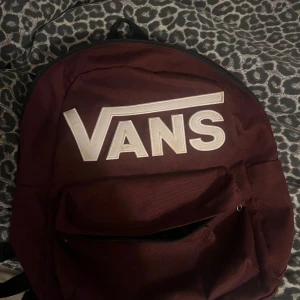 Vinröd ryggsäck från Vans - Snygg vinröd ryggsäck från Vans med stort vitt logotryck på framsidan. Väskan har ett stort huvudfack, en mindre ficka framtill med dragkedja och schackrutigt innerfoder. Perfekt för skolan eller häng med kompisar.