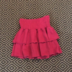 Rosa volangkjol med smock - Supersöt kort rosa kjol med bred smock i midjan och dubbla volanger. Kjolen har en luftig känsla och är perfekt för dig som gillar en lekfull och trendig stil. Materialet är mjukt och lätt, vilket gör den extra bekväm.