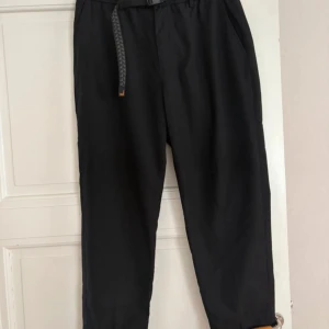 Svarta tapered byxor med bälte - Svarta byxor i tapered modell med elastisk midja och justerbart bälte med spänne. Tillverkade i 80% polyester och 20% viskos, vilket ger en mjuk och lätt känsla. Fickor i sidorna och bak, perfekt för en chill och stilren look.