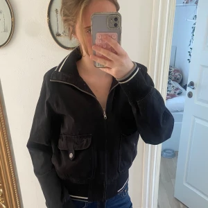 Svart bomberjacka från SIS by Sisters - Svart bomberjacka med dragkedja, två stora fickor med lock och knapp framtill samt ribbade muddar med vita ränder vid ärmslut och midja. Jackan är i bomull och nylon, har klassisk krage och sportig känsla.