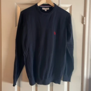Mörkblå tröja från Us polo assn - Klassisk mörkblå tröja från Us polo assn med rund halsringning och röd broderad logga på bröstet. Tröjan är långärmad och har ribbade muddar vid ärmslut och nederkant. Perfekt för en stilren och avslappnad look.