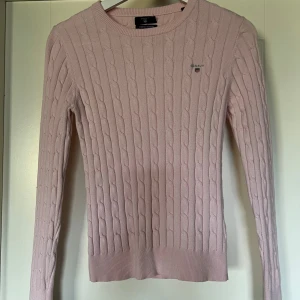 Ljusrosa kabelstickad tröja - Ljusrosa kabelstickad tröja från Gant i stretchig bomullsblandning. Klassisk rund halsringning, diskret logga på bröstet och ribbade muddar. Sista bilden visar hur färgen ser ut, de första bilderna stämmer inte riktigt i färgen. Använd och lite noppig men har mer att ge. 