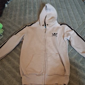 Beige Adidas hoodie med svarta ränder - Säljer en beige hoodie från Adidas med svarta ränder längs ärmarna och klassisk Adidas-logga på bröstet. Tröjan har huva, dragkedja framtill och två stora fickor. Perfekt för en sportig och avslappnad stil. Saknar snören och har blek märken på huvan