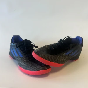 Svarta Adidas fotbollsskor med röd sula - Säljer ett par svarta Adidas fotbollsskor med coolt mönstrad ovandel och röda platta sulor. Skorna har blå detaljer och snörning framtill. Insidan är fodrad i blått och materialet är syntet för lätt känsla på planen. Perfekt för dig som vill sticka ut på träningen.