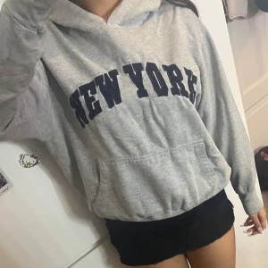 Grå hoodie med New York-tryck - Säljer en grå hoodie med svart New York-tryck över bröstet. Tröjan har huva, känguruficka och långa ärmar. Perfekt för chill dagar och streetstyle. Mjuk och skön, passar till det mesta.