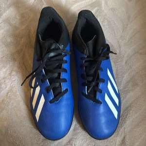Blå adidas fotbollsskor med dobbar - Säljer ett par blå adidas fotbollsskor med svarta snören och vit logga på sidorna. Skorna har en mönstrad sula med små dobbar för bra grepp på konstgräs. Snygg sportig look och klassisk adidas-design.