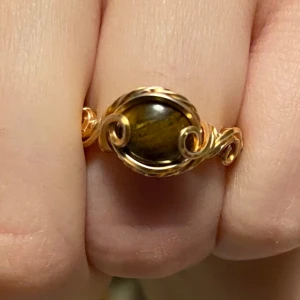 Guldfärgad ring med tigeröga  - Elegant guldfärgad ring med tigerögasten i mitten. Ringen har en vacker, snirklig design som ger ett unikt och stilrent intryck. Perfekt för att ge en touch av glamour till din look. - Nickelfri - Non-tarnish behandling - Storleksguide på sista bilden 