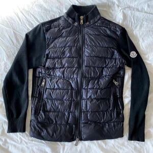 Moncler Cardigan S - Svart - Snygg svart cardigan från Moncler med quiltad front och stickade ärmar. Jackan har dragkedja framtill, ståkrage och två dragkedjeförsedda fickor. Klassisk Moncler-logga på ena ärmen och coolt serietryck på insidan. Perfekt för dig som vill ha en stilren och exklusiv look.