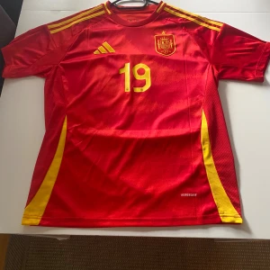 Spaniens landslagströja Lamine Yamal - Röd fotbollströja med gula detaljer, nummer 19 och Lamine Yamal på ryggen. Officiellt Spanien-emblem på bröstet och klassiska ränder på axlarna. Tillverkad i lätt och ventilerande material, perfekt för match eller träning.