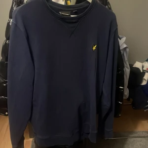 Mörkblå sweatshirt från Lyle & Scott - Säljer en mörkblå sweatshirt från Lyle & Scott med klassisk rund hals och gul logga på bröstet. Tröjan har långa ärmar och ribbade muddar vid hals, ärmslut och nederkant. Perfekt för dig som gillar stilren och enkel design.